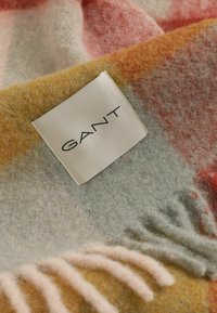Écharpe en laine douce avec un motif à carreaux jaune, rose et gris, bords frangés, et une étiquette rectangulaire blanche de la marque GANT cousue près d'un coin.
