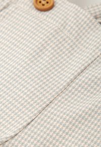 Tissu à carreaux bleu clair et crème avec un tissage texturé, comportant un bouton en bois et un détail de poche sur le vêtement.