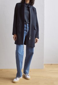 Marinblå zip-up-jacka med två framfickor, parat med ljusblå raka jeans och vita sneakers på ett trägolv.