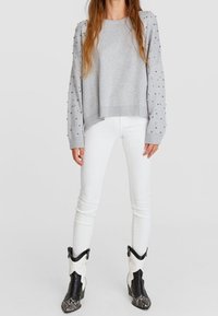 Sweat-shirt gris avec des clous décoratifs sur les manches, associé à un jean skinny blanc et des bottines noires de style western avec des accents blancs.