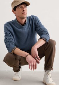 Un pull en tricot bleu avec un col en V, un pantalon brun et des baskets blanches montantes. La personne porte une casquette beige et a un bracelet marron.