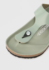 Πράσινη καουτσούκ σαγιονάρα με λουράκι για τα δάχτυλα και μαύρο κουμπί αξεσουάρ. Διαθέτει υφασμάτινη όψη στο  πέλμα και μεσαίο σόλο από ανοιχτό καφέ φελλό.