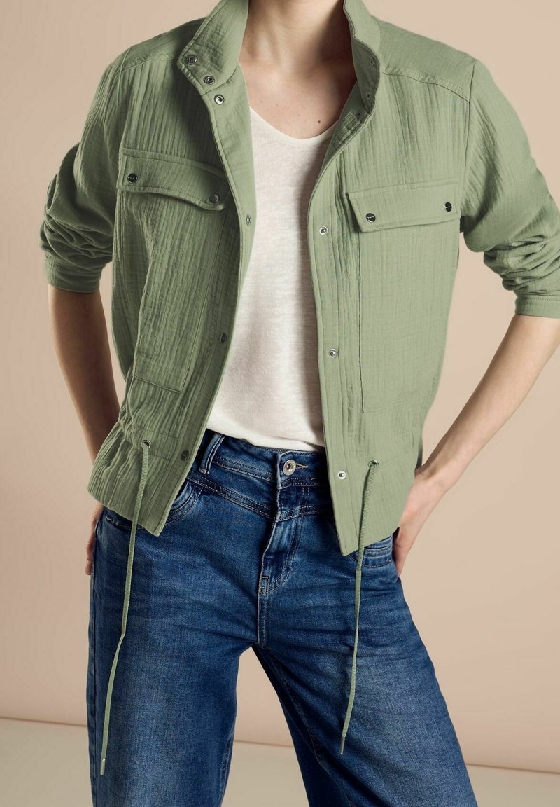 Personne portant une veste boutonnée vert clair sur un t-shirt blanc et un jean bleu, debout avec les mains sur les hanches devant un fond beige.