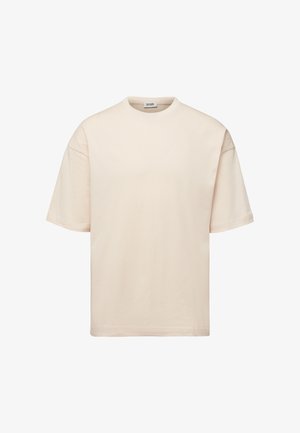 Beige lyhythihainen, väljämallinen puuvillainen T-paita pyöreällä pääntiellä ja näkyvällä merkkilapulla kauluksen sisäpuolella valkoista taustaa vasten.