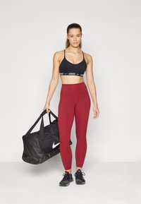 Črna športna modrc s belim pasom "Nike", visoko pasne burgundske legice in črne športne čevlje. Drži veliko črno torbo.