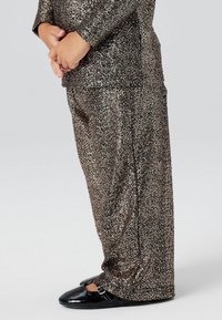 Glanzende bronzen outfit bestaande uit een top met lange mouwen en brede pijpen, gecombineerd met zwarte patenten schoenen. Zachte textuur met schitterende accenten.