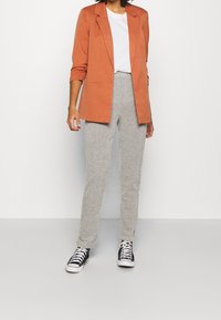 Pomarańczowa marynarka o dopasowanym kroju na białym T-shircie, zestawiona z jasnoszarymi teksturowanymi spodniami i czarno-białymi trampkami.