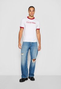 Bijela pamučna majica s crvenim rebrastim ovratnikom i rukavima, s logotipom "Calvin Klein". Plave traperice s izrezanim koljenima. Crne cipele s debelim đonom.