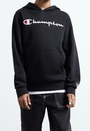 Person trägt einen schwarzen Champion-Hoodie mit Fronttasche, weißem Logo auf der Brust und schwarze Hose mit kontrastierenden Nähten.
