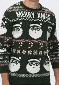 Maglione di Natale lavorato a maglia di colore verde scuro con volti di Babbo Natale bianchi con occhiali, bastoncini di zucchero, fiocchi di neve e il testo "MERRY XMAS" in un motivo festoso.