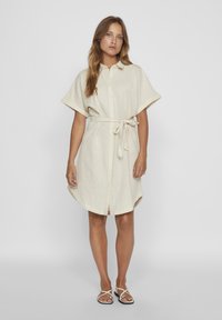 Bež haljina od lanenog platna s kratkim rukavima, dugmetima sprijeda i remenom na struku. Zaobljen rub i bočni prorezi poboljšavaju prozračnost.