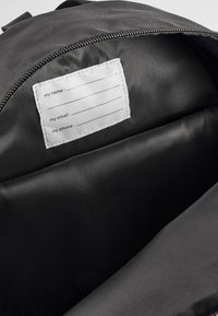 Intérieur d'un sac à dos noir avec un revêtement en tissu doux et une étiquette blanche comportant trois champs vides pour les coordonnées.