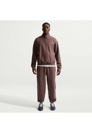 Mężczyzna stoi przodem, ubrany w brązowy dres z bluzą na ćwierćzamek, dopasowane spodnie, ciemne sneakersy oraz białą podkoszulkę, na jednolitym tle.