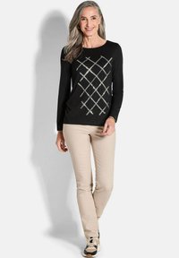 Schwarzes Langarmoberteil mit einem Diamantmuster und metallischen Akzenten, kombiniert mit beigen Hosen und schwarzen sowie beigen Sneakern.