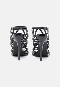 Sandálias pretas de salto alto com um design entrelaçado. Apresentam material brilhante de patente, tiras finas e um salto stiletto estreito.