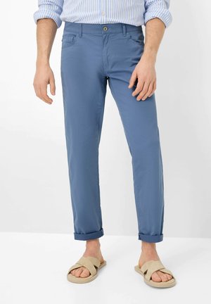 Homme portant un pantalon chino bleu retroussé, une chemise rayée et des sandales beiges, debout sur un fond blanc.