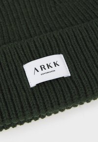 Tamnozeleni pleteni zimski šešir s rebrastom teksturom; ima bijelu tekstilnu oznaku s natpisom "ARKK COPENHAGEN" u crnom slovu.
