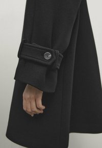 Sort uldfrakke med brede manchetter, der har en knap og syninger, der fremhæver en glat tekstur og skræddersyet design.