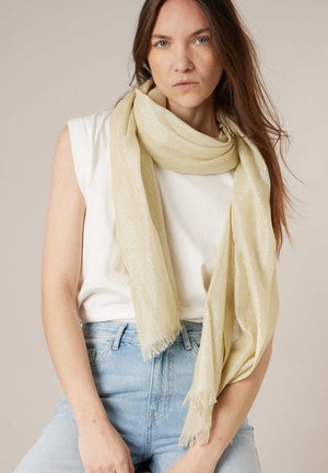 Jeune femme aux longs cheveux bruns porte un top blanc sans manches, un jean bleu clair et une écharpe crème scintillante drapée autour du cou.