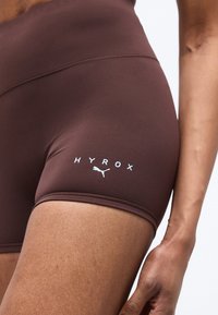 Εγγύς λήψη ενός ατόμου που φοράει καφέ αθλητικό σορτς με την επωνυμία Puma HYROX, εστιασμένο στην περιοχή του άνω μηρού και του γοφού.