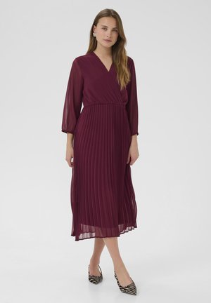 Abito plissettato bordeaux con scollatura a V e maniche a tre quarti, realizzato in materiale leggero, abbinato a scarpe a punta bianche e nere a righe.