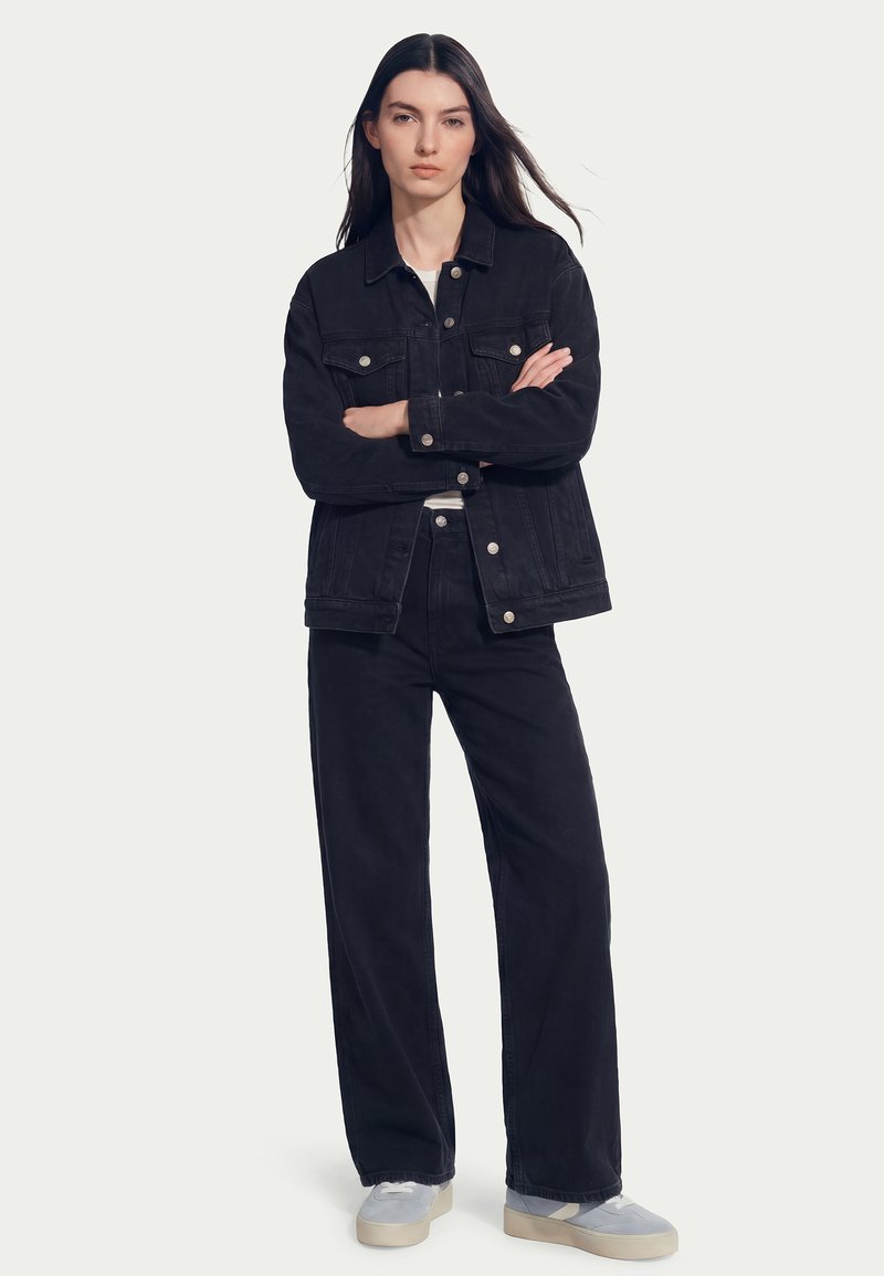 Jeune femme aux longs cheveux noirs portant une veste en denim noir et un pantalon assorti avec des baskets grises, debout les bras croisés sur un fond clair.