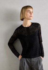 Čierny čipkový top s dlhým rukávom, zdobený zložitými geometrickými vzormi, okrúhlym výstrihom a rovný strih. V kombinácii so sivými elegantnými nohavicami.
