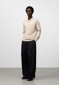 Maglione beige a maglia con colletto e dettaglio dei bottoni, abbinato a pantaloni neri a gamba larga e scarpe nere, su uno sfondo neutro.