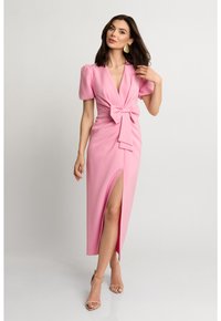 Różowa, dopasowana sukienka z głębokim dekoltem w kształcie litery V, krótkimi bufkami, detalem w postaci kokardy w talii, rozcięciem z boku i gładką fakturą. Połączona z nagimi sandałami na paski.