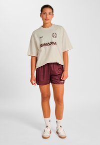 Lys beige oversized t-shirt med 'SWARM'-logo, kombineret med mørkerøde shorts. Model iført hvide sokker og atletikskor med brune detaljer.