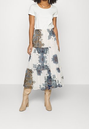 Vrouw draagt een wit topje met korte mouwen en een witte enkellange rok met blauwe en bruine paisley-patronen, gecombineerd met beige laarzen met hak.