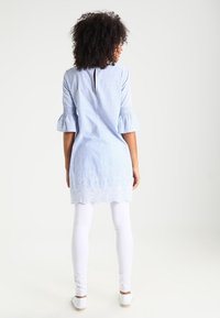 Tunique bleu clair à manches bouffantes, avec une ouverture au dos, un ourlet brodé, portée sur des leggings blancs. Tissu lisse et respirant.