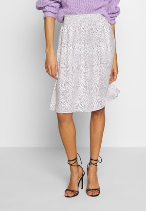 Kvinde iført en hvid knælængde nederdel med sorte prikker, en lavendelfarvet strikket sweater og sorte sandaler med hæl og stropper.