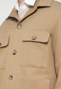 Μπεζ σακάκι κατασκευασμένο από υφασμάτινη υφή, με δύο τσέπες στο στήθος και στρογγυλά καφέ κουμπιά. Το σχέδιο περιλαμβάνει γιακά και ορατές διακοσμητικές ραφές.