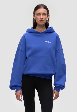 Blauer Hoodie mit einer Vordertasche und gerippten Bündchen. Mit einem weißen Logo auf der Brust. Kombiniert mit schwarzen Leggings. Glatte Textur.