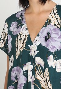 Kukkamallinen lyhythihainen mekko tummanvihreänä, jossa on suuria laventelin- ja valkoisia kukkia, syvä V-kaulus ja pehmeä, sileä kangas.