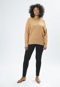 Brun striktrøje med ribbet kant, afslappet pasform og detaljer i siderne, båret med sorte leggings og brune mule-sko med guld accenter.