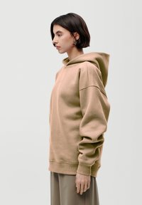 Felpa oversize beige con polsini e orlo a costine, texture morbida, dotata di cappuccio con cordino. Il modello è in profilo contro uno sfondo neutro.
