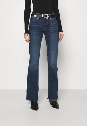 Frau trägt dunkelblaue Bootcut-Jeans mit einem schwarzen Gürtel, kombiniert mit schwarzen Stiefeletten mit Absatz und einem schwarzen Langarmshirt.