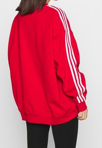 Röd oversized sweatshirt med vit tre-stripes design längs ärmarna, tillverkad av mjukt tyg. Har rund halslinning och ribbade ärmslut.