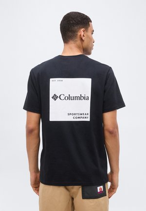 Άνδρας που φοράει μαύρο T-shirt Columbia με μεγάλο λευκό λογότυπο σε τετράγωνο στο πίσω μέρος, μπεζ σορτς με μαύρο patch στην τσέπη, στάθηκε μπροστά σε λευκό φόντο.