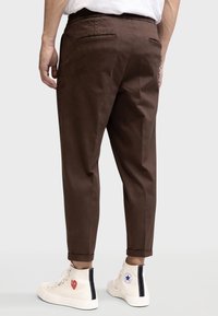 Pantalons taillés marron avec une jambe légèrement fuselée, poches arrière, et ourlets retroussés, associés à des baskets montantes blanches avec des graphismes.
