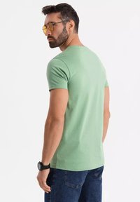 T-shirt in cotone verde con maniche corte, scollatura rotonda e una texture liscia. Indossato con jeans blu scuro e un orologio nero.