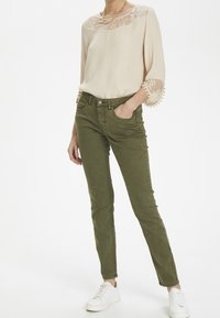 Ljus beige blus med spetsdetaljer och pom-pom detaljer, ihop med slim fit olivgröna byxor och vita sneakers. Slät textur, avslappnad passform.