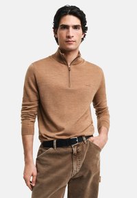 Suéter marrón con cuello tipo cremallera en una textura suave, de mangas largas, combinado con pantalones caqui y un cinturón negro. Marca sutil cerca del cuello.