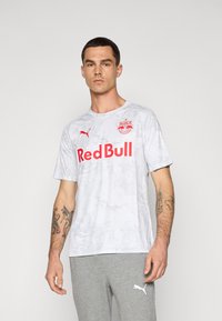White camo-patterned short-sleeve shirt with red "Red Bull" text and logo. Made of a lightweight material; worn with grey sweatpants.