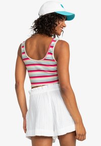 Gestreepte crop top in levendige kleuren met zigzagpatroon; gecombineerd met witte, gestructureerde shorts met een elastische tailleband. Witte pet met blauwe accenten.