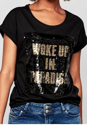 Czarna koszulka z napisem z złotych cekinów „"Woke Up In Paradise"”, noszona z niebieskimi dżinsami.