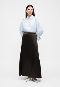 Femme portant une chemise bleu clair oversize, une jupe longue noire taille haute et des chaussures blanches pointues, debout sur un fond blanc.