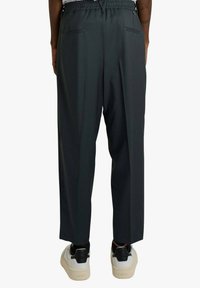 Vista posteriore di una persona che indossa pantaloni larghi a gamba larga con vita elastica nera e scarpe da ginnastica bianche con dettagli neri, in piedi su uno sfondo semplice.